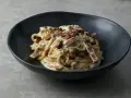 Pyszne spaghetti carbonara, jeden z klasycznych makaronów, podane w ciemnej misce, posypane natką pietruszki i kawałkami boczku.