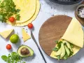 Przygotowanie keto tortilli z awokado, rukolą i serem. Na stole świeże pomidorki, limonka i przyprawy.