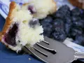 Wilgotne ciasto z borówkami, z widocznymi owocami w środku, czeka na widelcu. Obok świeże borówki.