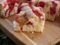 Puszyste ciasto na kefirze z rabarbarem i truskawkami, posypane cukrem pudrem, na drewnianej desce.