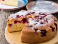 Wilgotne ciasto jogurtowe z owocami, posypane cukrem pudrem, wygląda apetycznie. Kawałek ciasta na talerzu obok.