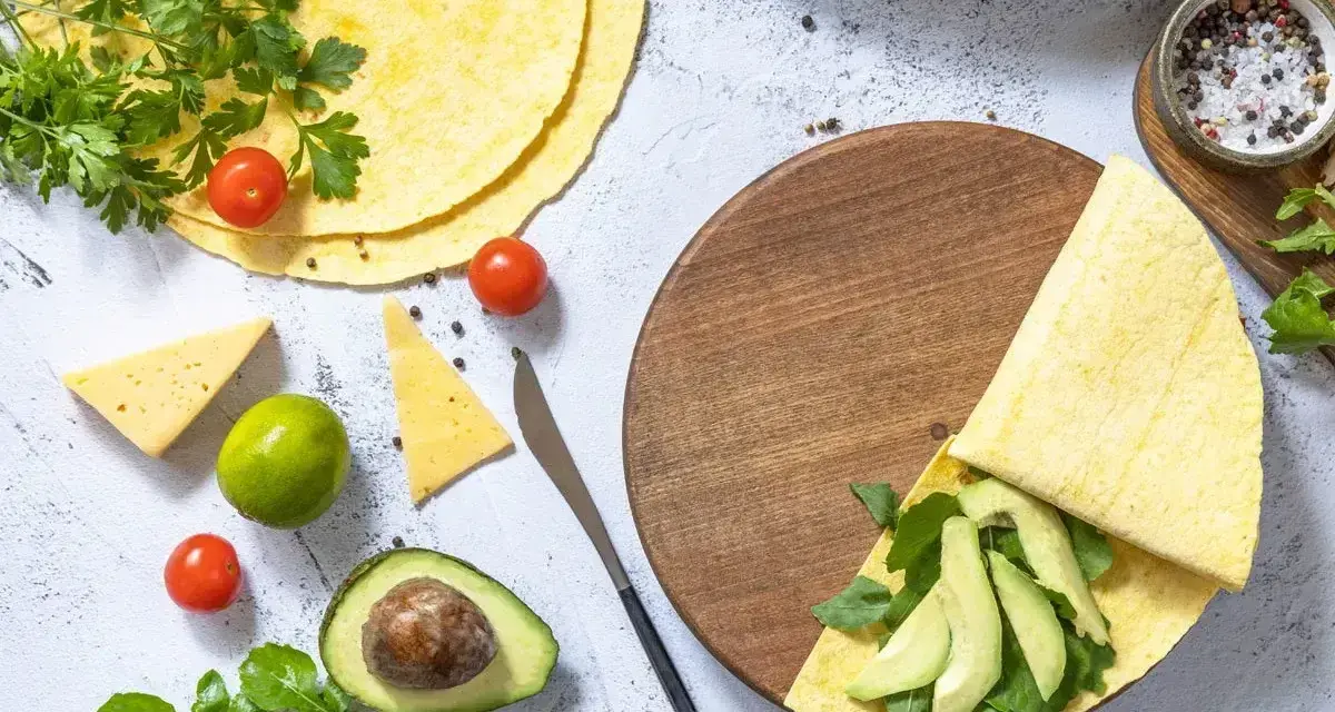 Przygotowanie keto tortilli z awokado, rukolą i serem. Na stole świeże pomidorki, limonka i przyprawy.