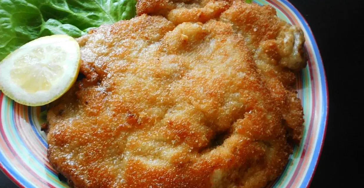 Złocisty kotlet schabowy Magdy Gessler, podany z liściem sałaty i plasterkiem cytryny na kolorowym talerzu.