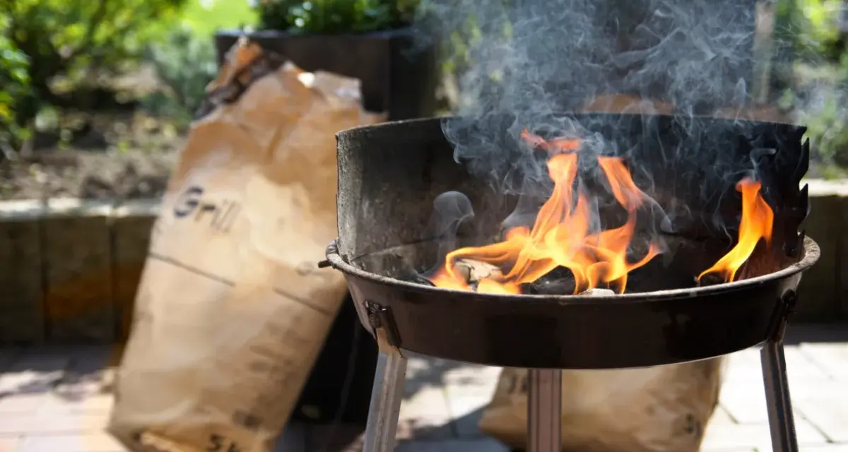 Rozpalony grill z płomieniami i dymem, obok torba z węglem. Idealny początek, by dowiedzieć się, jak rozpalić grilla.