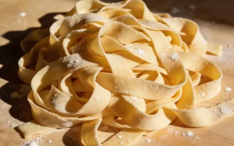 Świeży, domowy makaron tagliatelle, gotowy do zupy z makaronem. Złociste wstążki mączne, lekko oprószone mąką, ułożone na drewnianej desce.