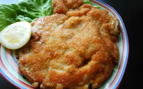 Złocisty kotlet schabowy Magdy Gessler, podany z liściem sałaty i plasterkiem cytryny na kolorowym talerzu.
