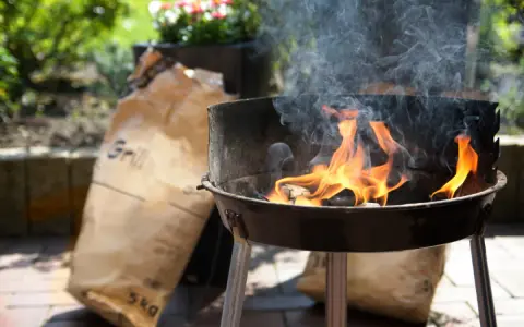 Rozpalony grill z płomieniami i dymem, obok torba z węglem. Idealny początek, by dowiedzieć się, jak rozpalić grilla.