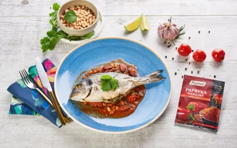 Dorada ryba w sosie pomidorowo-fasolowym, udekorowana kolendrą. Obok miseczka fasoli, czosnek, limonka i pomidorki.
