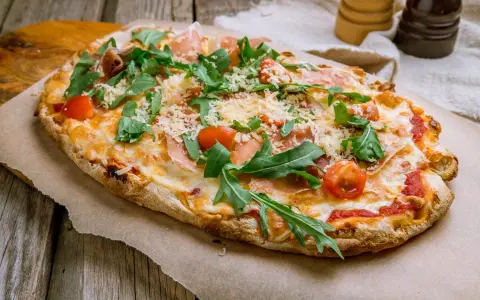 Pinsa co to? Aromatyczna, podłużna pizza z rukolą, pomidorkami i szynką parmeńską, posypana parmezanem.