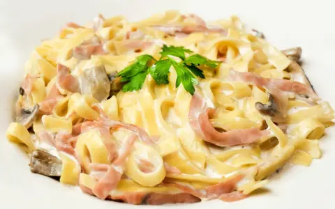 Tagliatelle z szynką i pieczarkami w kremowym sosie, z dodatkiem mozzarelli. Udekorowane natką pietruszki.