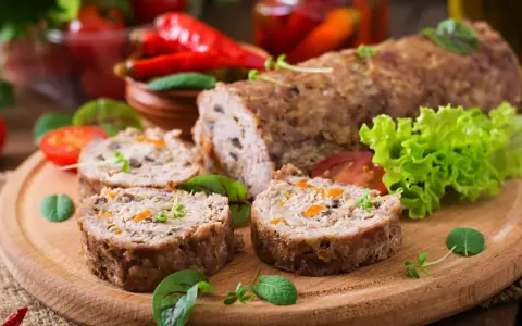 Pokrojone zrazy z mięsa mielonego z warzywami na desce, obok sałata, pomidory i papryczki chili.