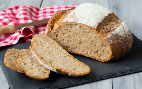 Pyszny, świeżo upieczony chleb pełnoziarnisty z chrupiącą skórką, pokrojony na kromki, leży na czarnej desce. Obok czerwono-biała ściereczka i nóż.