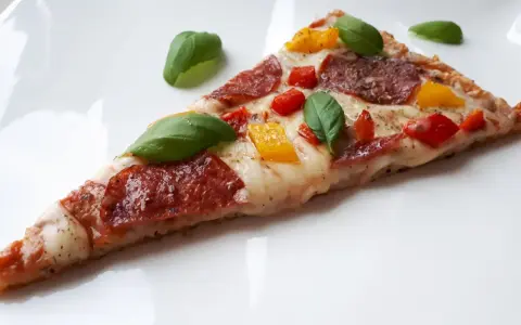 Kawałek pysznej pizzy keto z salami, papryką i bazylią na białym talerzu.