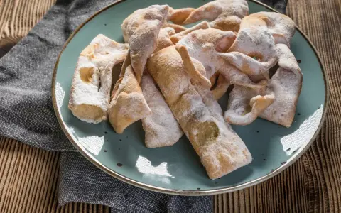 Chrupiące faworki, przygotowane według przepisu babci, posypane cukrem pudrem, leżą na talerzu.