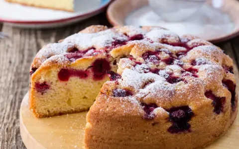 Wilgotne ciasto jogurtowe z owocami, posypane cukrem pudrem, wygląda apetycznie. Kawałek ciasta na talerzu obok.