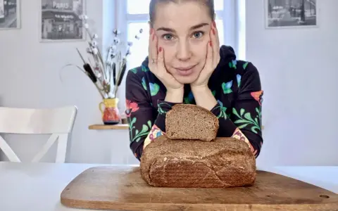 Kobieta z dłońmi podpartymi pod brodą patrzy na pyszny chleb wieloziarnisty. Jeden bochenek leży na drugim, na drewnianej desce.