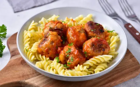 Pulpety w sosie pomidorowym jak u babci, podane z makaronem fusilli. Danie ozdobione posiekaną natką pietruszki.