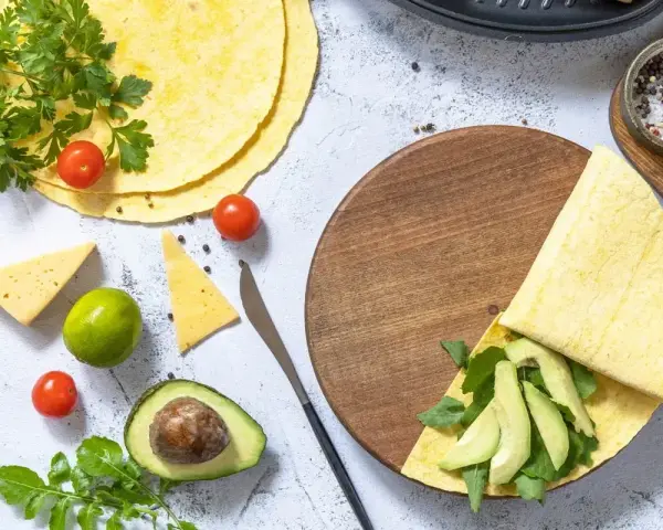 Przygotowanie keto tortilli z awokado, rukolą i serem. Na stole świeże pomidorki, limonka i przyprawy.