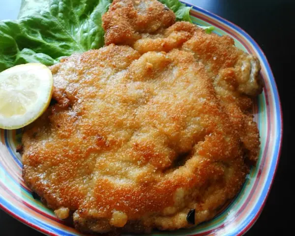Złocisty kotlet schabowy Magdy Gessler, podany z liściem sałaty i plasterkiem cytryny na kolorowym talerzu.