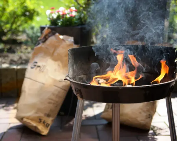 Rozpalony grill z płomieniami i dymem, obok torba z węglem. Idealny początek, by dowiedzieć się, jak rozpalić grilla.