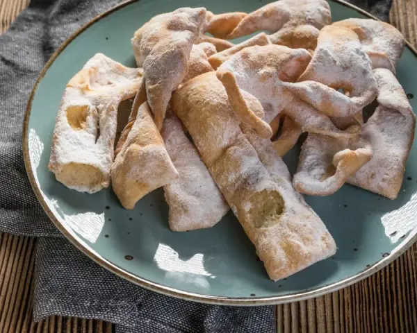 Chrupiące faworki, przygotowane według przepisu babci, posypane cukrem pudrem, leżą na talerzu.