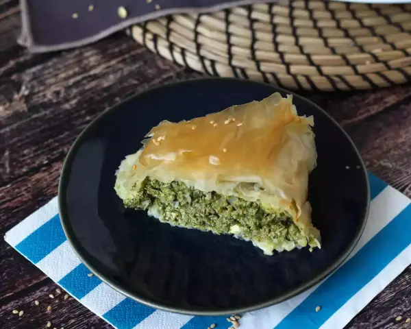 Kawałek spanakopity z chrupiącym ciastem filo i zielonym nadzieniem ze szpinaku i sera feta, posypany sezamem.