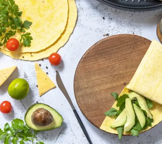 Przygotowanie keto tortilli z awokado, rukolą i serem. Na stole świeże pomidorki, limonka i przyprawy.