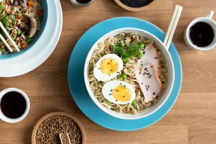 Miseczka tantanmen z makaronem, jajkiem, plastrami mięsa i zielonym koperkiem, obok sos sojowy i przyprawy.