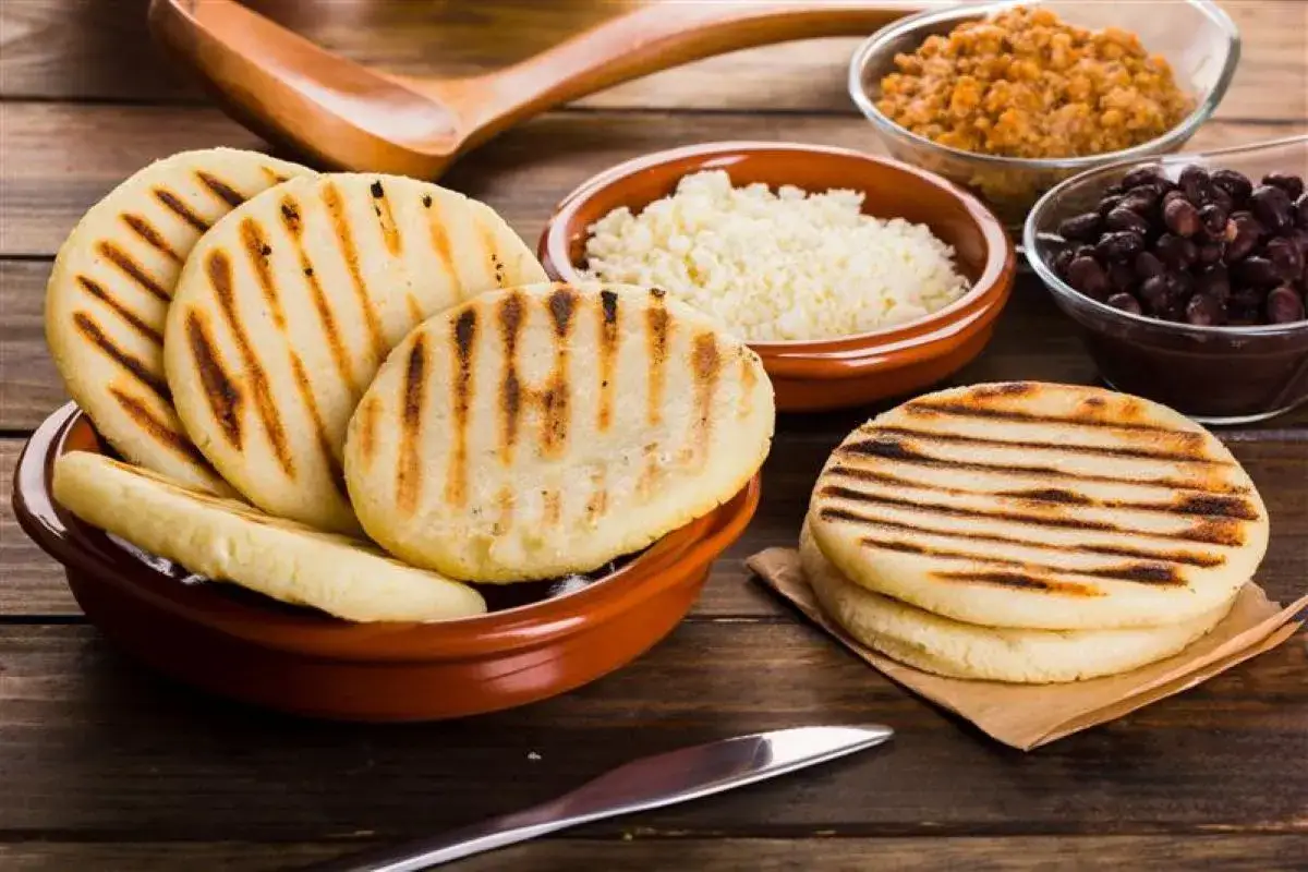 Grillowane arepas w ceramicznej misce, obok miseczki z białym serem, czarną fasolą i brązowym gulaszem.