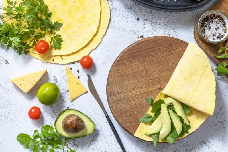 Przygotowanie keto tortilli z awokado, rukolą i serem. Na stole świeże pomidorki, limonka i przyprawy.